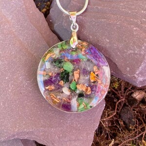 Può includere: Una collana con un ciondolo rotondo e trasparente su una catena d'argento. Il ciondolo è pieno di una varietà di piccole pietre colorate, tra cui ametista, aventurina verde e scaglie di rame.