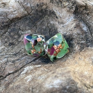 May include: A pair of square-shaped earrings with green, pink, and gold flecks embedded in clear resin.