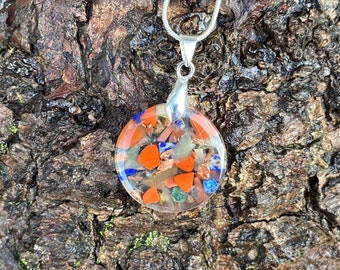 May include: A round, clear pendant necklace with a silver chain. The pendant is filled with small, colorful stones, including red, orange, blue, green, and brown.