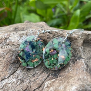 May include: A pair of silver earrings with oval-shaped green and pink gemstone resin pieces. The resin is clear and shows the gemstones and gold flecks inside.