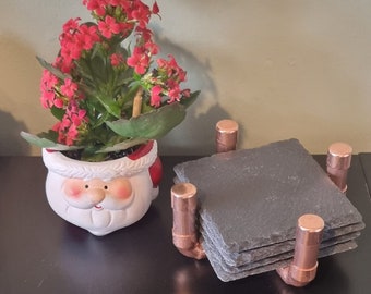May include: A set of four black slate coasters with copper pipe accents. The coasters are stacked on top of each other and are sitting on a black surface. A small potted plant with red flowers is in the background.