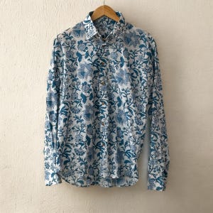 Könnte beinhalten: Ein langärmeliges Hemd mit Knopfleiste auf weißem Grund und einem sich wiederholenden blauen Blumen- und Pflanzenmuster. Das Kleidungsstück verfügt über einen klassischen Kragen, eine Knopfleiste mit braunen Knöpfen und lange Ärmel mit Manschetten.