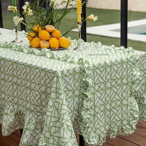 Könnte beinhalten: Eine weiße Tischdecke mit grünem geometrischem Gittermuster und gerüschten Kanten. Der Stoff liegt auf einem Tisch, darauf steht eine Schale mit gelben Zitronen und weißen Blumen in einer silbernen Vase. Der Text Jaipur Store Studio ist sichtbar.