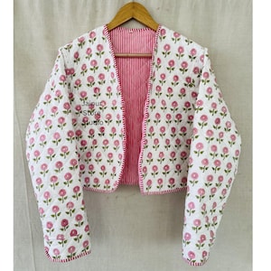 Könnte beinhalten: Weiß gesteppte Jacke mit rosa Blumenmuster und rot-weiß gestreiftem Futter. Die Jacke ist offen und zeigt das Innenfutter. Der Text "Jaipur Store Studio" ist sichtbar.