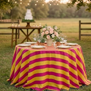 May include: An outdoor dining table with a vibrant tablecloth in yellow and magenta stripes. The table is set with white plates, wine glasses, and a floral arrangement. A tiered cake sits on a nearby table.
