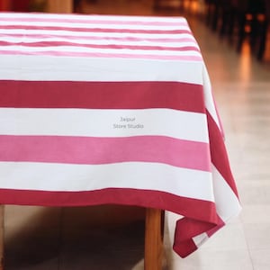 May include: A striped tablecloth in shades of red, pink, and white covers a wooden table. The fabric features broad horizontal stripes. The text "Jaipur Store Studio" is printed on the tablecloth.