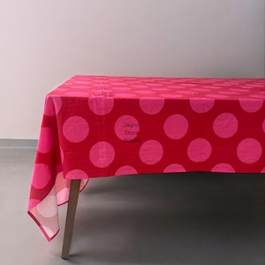 Puede incluir: Un mantel rojo con lunares rosas grandes cubre una mesa de madera. El mantel tiene el texto "Jaipur Store Studio" impreso. La tela está drapeada sobre la mesa, mostrando el patrón.