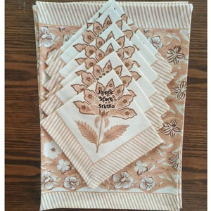Peut inclure: Un ensemble de serviettes florales crème et une nappe assortie. Les serviettes présentent un grand motif floral dans des tons bruns et sont bordées d'une bordure rayée. La nappe a un motif floral et des bordures rayées. Le texte "Jaipur Store Studio" est visible.