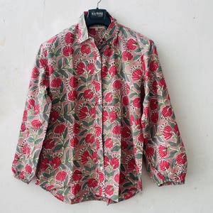 May include: A long-sleeved button-down shirt in a muted pink hue featuring a repeating floral pattern of vibrant red blossoms and sage green leaves. The garment includes a classic collar, a front button placket, and gathered cuffs at the wrists.