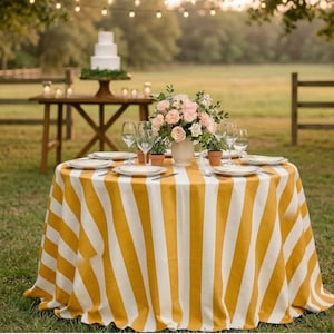 May include: A round table covered with a mustard yellow and white vertical striped tablecloth. The table is set with white plates, clear glassware, and a central floral arrangement featuring pink roses in a light-colored vase.