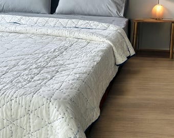 May include: A white quilted bedspread with a geometric pattern and blue trim is draped over a bed. The bed has gray pillows and a blue headboard. A wooden side table with a lamp is next to the bed.