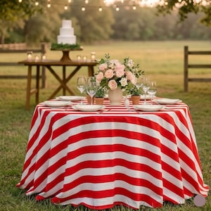 Può includere: Un tavolo rotondo coperto da una tovaglia a righe rosse e bianche, apparecchiato con piatti bianchi, bicchieri trasparenti e un centrotavola floreale di rose rosa e bianche in un vaso bianco, posizionato all'aperto su un prato.