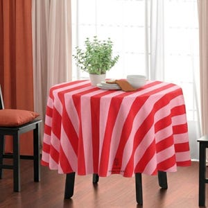 May include: A round table covered with a tablecloth featuring alternating vertical stripes in red and light pink. On the table sits a small potted green plant in a white pot, a stack of white plates, a white bowl, and a folded brown napkin.