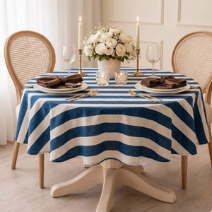 May include: A round table covered with a blue and white striped tablecloth, set with two white plates, brown napkins, gold forks, wine glasses, two tall candles in gold holders, two small glass votive candle holders, and a white floral centerpiece.