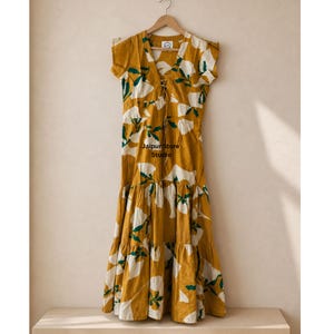 Könnte beinhalten: Senfgelbes Maxikleid mit einem Blumenmuster in Weiß und Grün. Das Kleid hat einen V-Ausschnitt mit Schnürdetail, kurze Ärmel und einen Stufenrock. Der Text "Jaipur Store Studio" ist auf dem Kleid zu sehen.