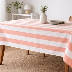 Pode incluir: Uma toalha de mesa retangular com listras largas alternadas brancas e coral. A toalha de mesa está sobre uma mesa de madeira. Um vaso com vegetação, uma tigela e pratos estão sobre a mesa. A mesa está sobre um tapete tecido.