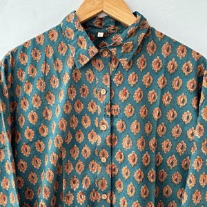 Puede incluir: Camisa abotonada de color turquesa con un estampado floral repetido en naranja y marrón. La camisa tiene cuello y botones de madera. La tela parece ligera, adecuada para climas cálidos.