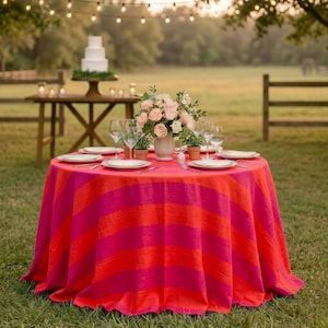 Puede incluir: Una mesa redonda preparada para un evento al aire libre, cubierta con un mantel a rayas rosa y naranja vibrante. La mesa está puesta con platos blancos, copas de vino y un centro de mesa floral. Una tarta de varios pisos se encuentra sobre una mesa de madera en el fondo.