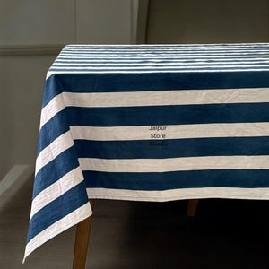 May include: A rectangular tablecloth with alternating wide stripes of navy blue and white. The tablecloth is draped over a wooden table, with the stripes running horizontally across the surface. The text "Jaipur Store Studio" is visible on the tablecloth.