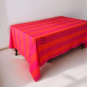 May include: A rectangular tablecloth with horizontal stripes in shades of pink and orange. The tablecloth is draped over a table, with the text "Jaipur Store Studio" visible on the fabric.