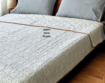 May include: A white quilted bedspread with a brown border is neatly arranged on a wooden bed frame. Two gray pillows rest on the bed. The text "Jaipur store Studio" is visible on the bedspread.
