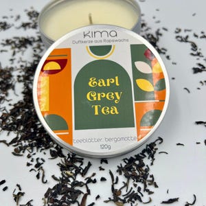 Könnte beinhalten: Eine silberne Dose mit einer weißen Kerze, auf der "Kima Duftkerze aus Rapswachs Earl Grey Tea Teeblätter Bergamotte 120g" steht. Die Kerze ist von losen schwarzen Teeblättern umgeben.