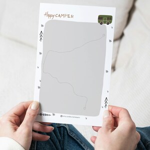 Può includere: Una carta bianca con la scritta "Happy Camper" in marrone. La carta presenta un disegno di mappa grigia, un'illustrazione di camper verde e piccole illustrazioni di alberi e foglie. La carta è tenuta da una persona che indossa jeans blu.