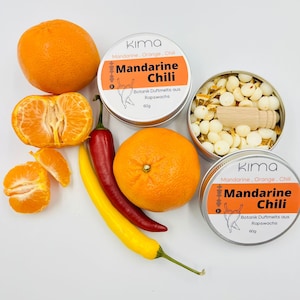 Könnte beinhalten: Draufsicht mit zwei Dosen "Mandarine Chili" Duftwachs, Orangen und Chilischoten. Eine Dose ist geöffnet und zeigt weiße Wachs-Melts und einen kleinen Holzschaber. Die andere Dose ist geschlossen. Auf weißem Hintergrund.