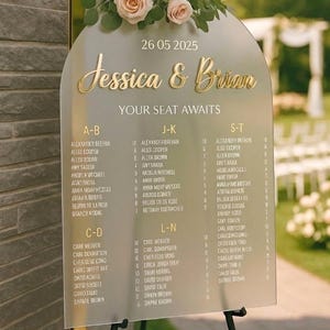 May include: An arched frosted acrylic wedding seating chart sign with gold lettering reading Jessica & Brian, 26 05 2025, and YOUR SEAT AWAITS. The sign is topped with a floral arrangement of three pale pink roses and green eucalyptus leaves.