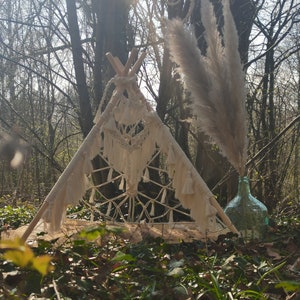 Op de afbeelding: Een witte macramé tipi tent met franjes die aan de randen hangen, opgesteld in een bos met hoge bomen en een glazen fles met gedroogd pampasgras.