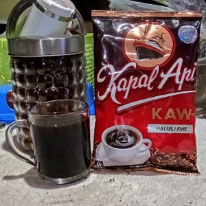 Puede incluir: Una taza de cristal transparente llena de café negro junto a una bolsa roja y marrón de café Kapal Api Kaw Halus. La bolsa muestra una ilustración de un barco, el texto Kapal Api, KAW, HALUS / FINE y una imagen de una taza blanca.