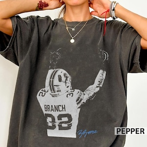 May include: A charcoal gray short-sleeve t-shirt featuring a distressed white graphic of a football player wearing a helmet and jersey with the name BRANCH and the number 32 on the back, with both arms raised in a celebratory gesture.