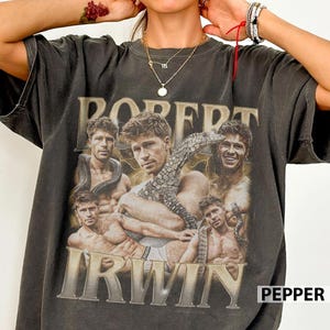 May include: A charcoal grey short-sleeve t-shirt featuring a vintage-style graphic print of Robert Irwin. The design includes multiple shirtless portraits of the individual, a lizard, and yellow lightning accents with the text ROBERT IRWIN.