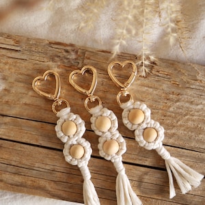 Puede incluir: Tres llaveros con anillos de metal dorado en forma de corazón y borlas de macramé blanco. Cada llavero tiene dos cuentas de madera.