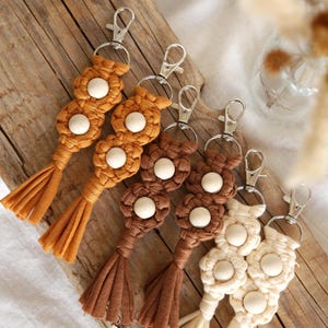 Peut inclure: Porte-clés en macramé dans les tons orange, marron et crème. Chaque porte-clés présente un motif de fleur tissée avec un centre de perle en bois et une frange. Les porte-clés sont attachés à des fermoirs métalliques argentés.