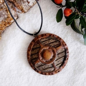 Op de afbeelding: Een houten schild hanger ketting met een donkerbruin leren koord. Het schild is gegraveerd met een gedetailleerd ontwerp en heeft een ronde vorm.