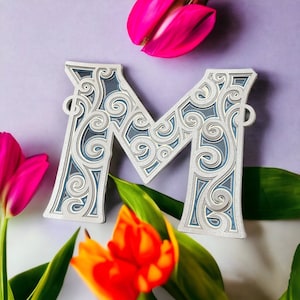 May include: A decorative, silver-colored letter M with a blue and white intricate design. The letter is surrounded by pink and orange tulips and green leaves.