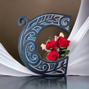 May include: A blue and silver decorative letter C with intricate floral designs. The letter is standing on a white fabric surface with a bouquet of red roses in front of it.