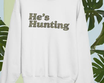 May include: White sweatshirt with the text "He's Hunting" in a camouflage pattern. The sweatshirt is displayed on a hanger against a backdrop of green and blue foliage.