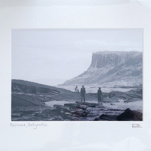 Può includere: Fotografia in bianco e nero di una costa rocciosa con una grande scogliera sullo sfondo. Quattro persone sono in piedi sulla spiaggia in primo piano. La didascalia recita "Fairhead, Ballycastle".