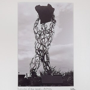 Può includere: Fotografia in bianco e nero di una scultura in metallo di una persona che solleva un grande masso. La scultura è realizzata con pezzi di metallo intrecciati e si trova all'aperto. Il testo "Protector of the Lough, Antrim" è scritto sotto l'immagine.