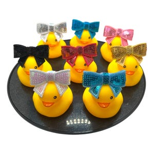 May include: Eight yellow rubber duck toys with different coloured sequin bows. The bows are black, white, pink, red, blue, gold, and two shades of blue.
