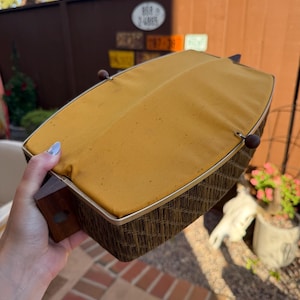 May include: A rectangular sewing basket featuring a dark brown woven wicker base and a mustard yellow fabric lid. The lid is secured with a thin metal frame and two small dark brown wooden knobs on the sides, with a wooden handle attached.