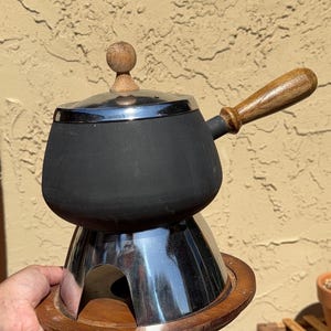 May include: A black fondue pot with a chrome lid and a wooden handle, featuring a matching wooden knob on top. The pot rests on a polished chrome stand, which is set upon a circular wooden base, creating a complete set for serving warm food.