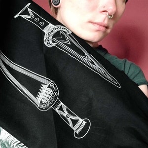 Puede incluir: Bufanda de tela negra con dos diseños de puñales estilizados blancos. Un puñal tiene un mango circular con un patrón repetitivo. El otro puñal tiene un mango más puntiagudo con un patrón repetitivo.