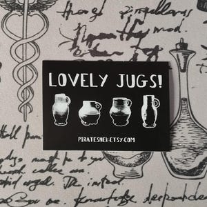 Peut inclure: Un autocollant rectangulaire noir avec le texte "LOVELY JUGS!" et l'adresse du site Web "PIRATESNEK.ETSY.COM". L'autocollant présente quatre illustrations blanches de cruches. Le fond est un tissu gris clair avec des écritures et des illustrations noires.