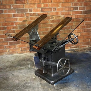 May include: A vintage industrial drafting table with a glass top and wooden supports. The table has a black metal base with a crank and a wheel for adjusting the height and angle of the top.