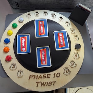 Könnte beinhalten: Ein Holzspielbrett mit einer schwarzen Mitte und einem drehbaren Zifferblatt. Das Zifferblatt hat die Zahlen 1 bis 10 und das Wort "Twist" darauf gedruckt. Auf dem Brett liegen vier Karten mit der Aufschrift "Phase 10".