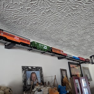 May include: A model train set with an orange and brown locomotive and several boxcars, mounted on a track and displayed on a white wall.