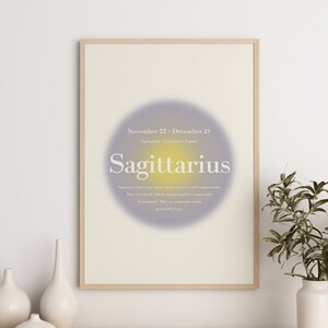 May include: A framed print with a purple and yellow gradient background. The text "November 22 - December 21" and "Sagittarius" is printed in white. The print is on a wooden shelf with two white vases and a diffuser.